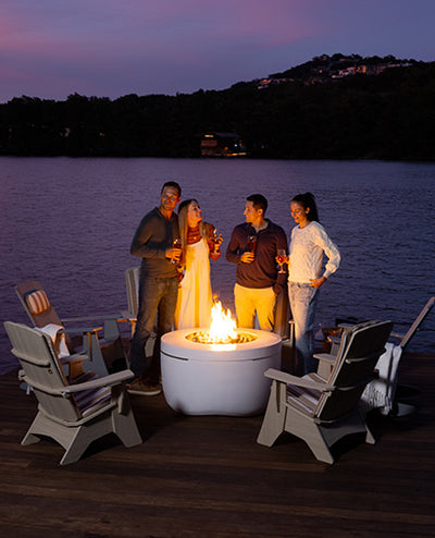 A group of four people stand around a glowing fire pit on a wooden deck by the water at dusk. They are holding drinks and appear to be enjoying the evening. Empty chairs surround the fire pit, and the sky is a deep blue with hints of purple.