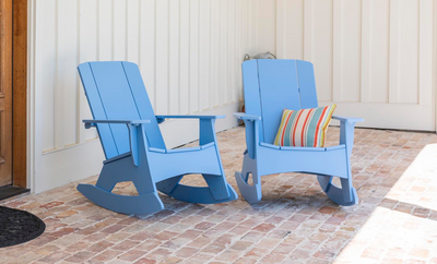 Two blue wooden rocking chairs sit on a brick patio near a white wall. One chair has a colorful striped pillow on it, while the other is empty. Sunlight casts soft shadows on the ground.